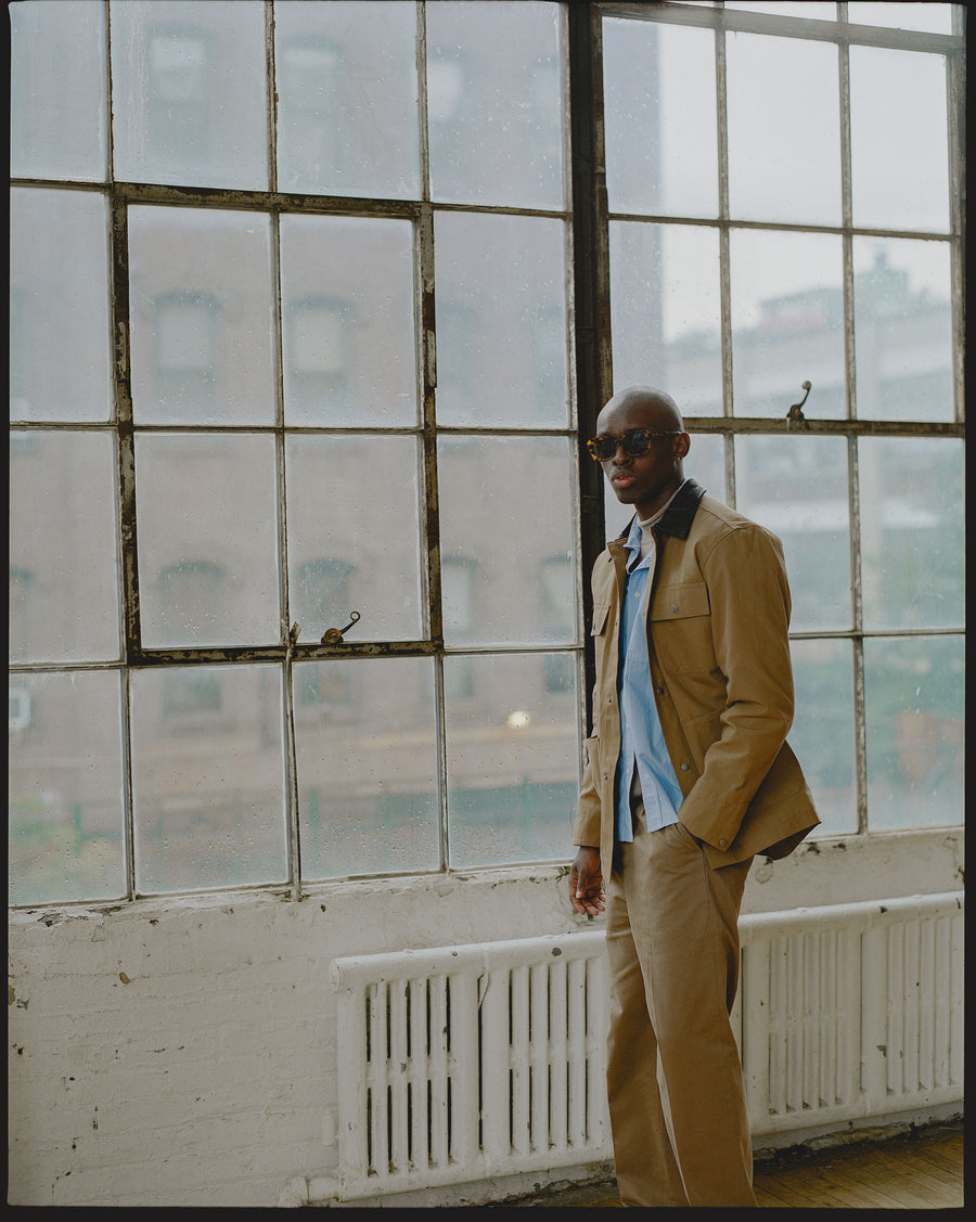 A man in a tan jacket and blue striped button down shirt stands in front of big windows.