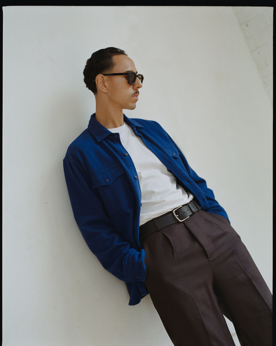 A man wearing sunglasses and an electric blue shirt leans against a wall.
