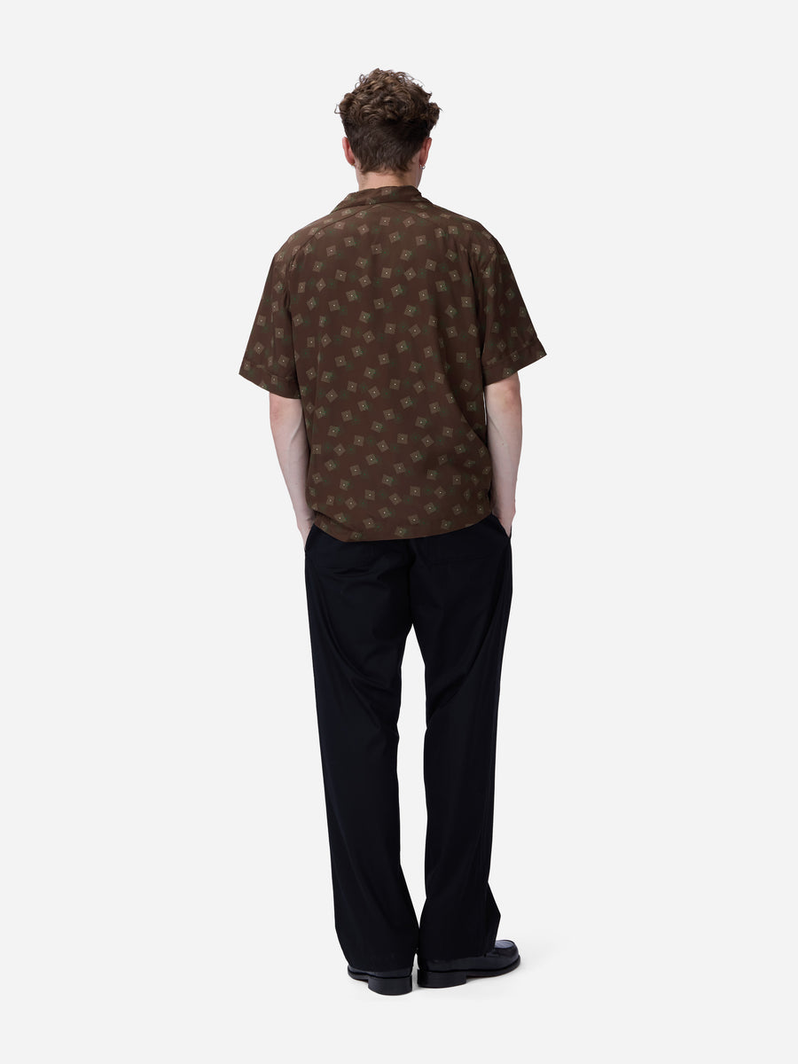 A man viewed from behind wearing a brown short-sleeve button-up shirt with a repeating circular monogram pattern, paired with black trousers and black shoes, against a light gray studio background.