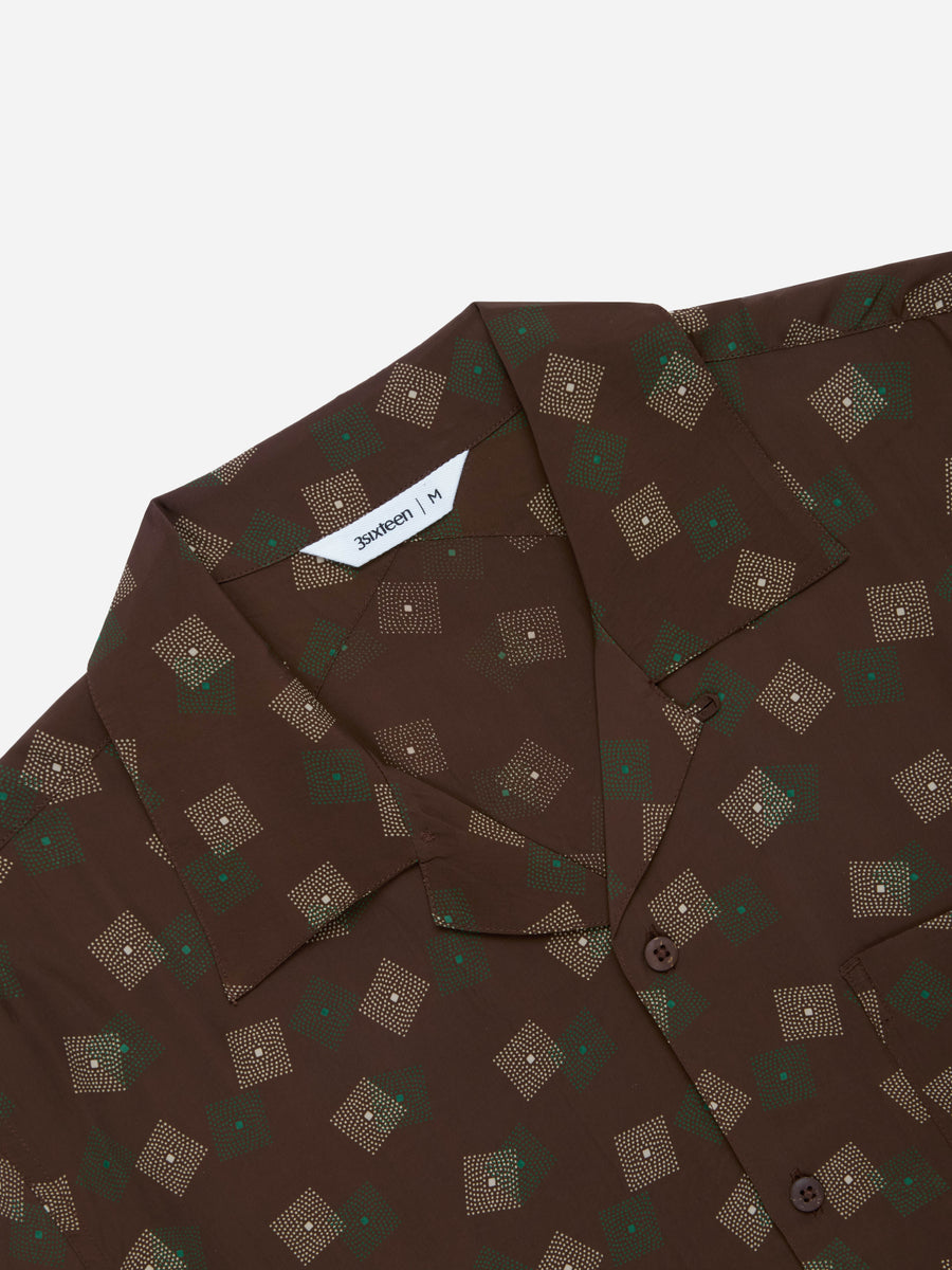 A close-up of a brown short-sleeve button-up shirt with a repeating geometric pattern of small squares and dots in beige, green, and white. The brand name 'Benedict' is visible on the inner neck label.