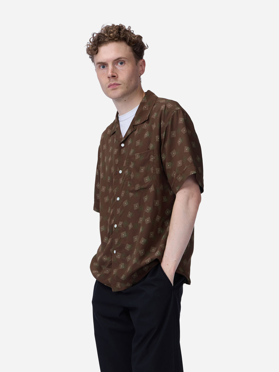 A man wearing a brown short-sleeve button-up shirt with a repeating beige monogram pattern, styled over a white undershirt and black trousers, photographed against a light gray studio background.