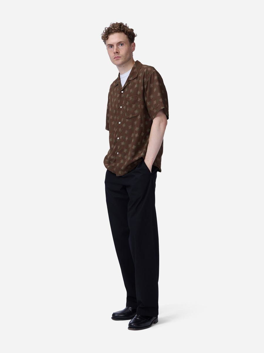 A man modeling a brown short-sleeve button-up shirt with a small repeating geometric pattern, paired with black trousers and black shoes, against a light gray studio background.
