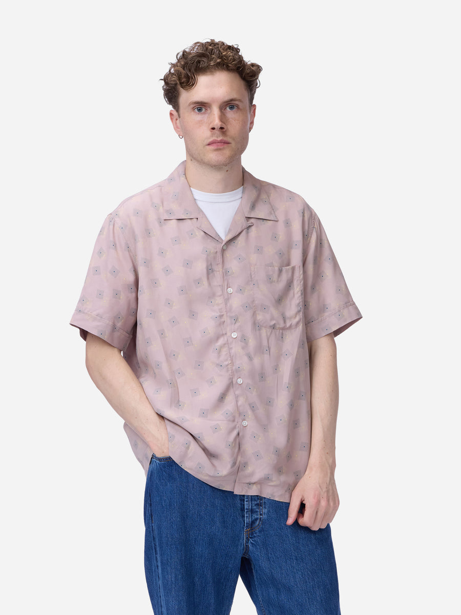 A man wearing a light pink short-sleeve button-up shirt with a subtle floral pattern, layered over a white t-shirt and blue jeans, against a plain light gray background.