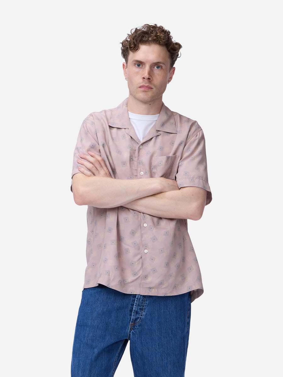 A man with curly brown hair wearing a light pink short-sleeve button-up shirt with a subtle floral pattern, layered over a white t-shirt and blue jeans, standing against a solid light gray background.