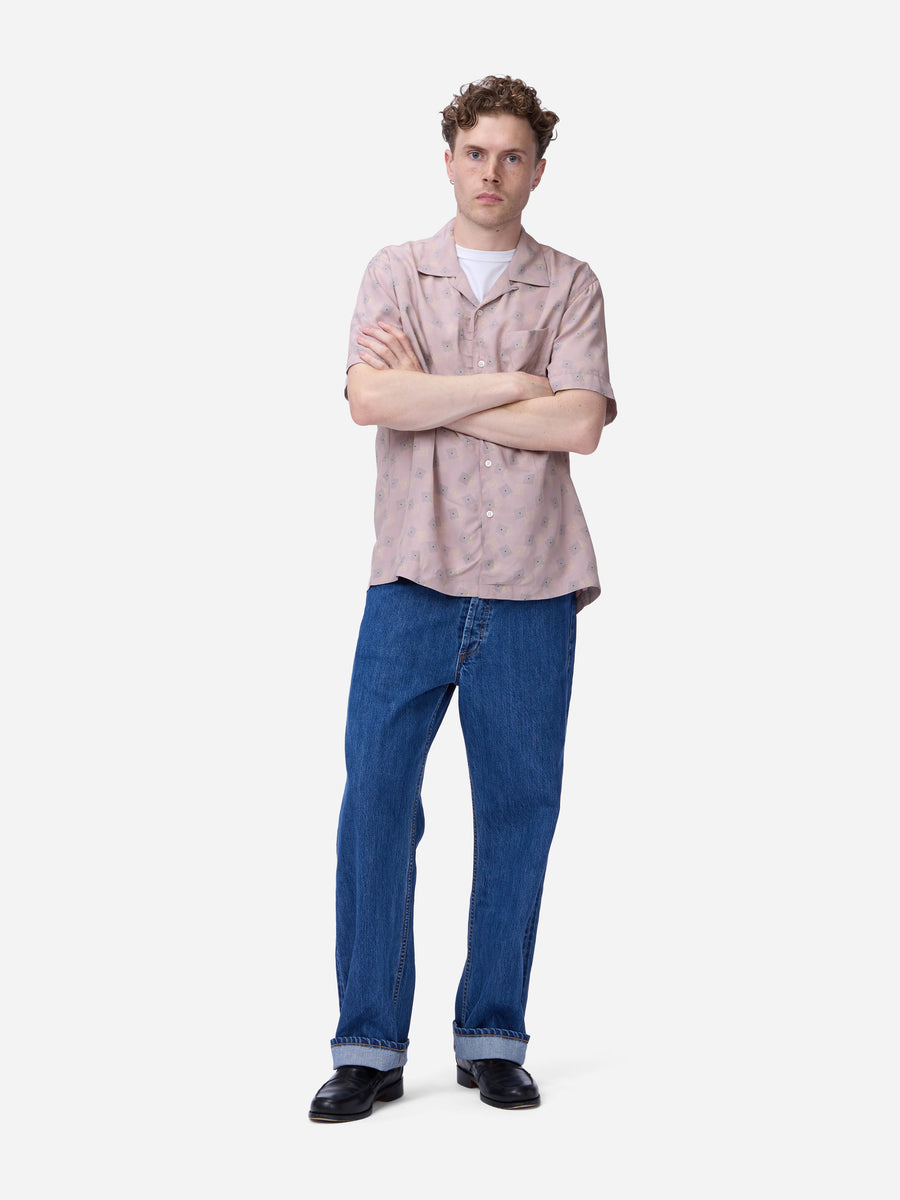 A man stands with arms crossed wearing a light pink short-sleeve button-up shirt, blue jeans, and black shoes against a plain light gray studio background.