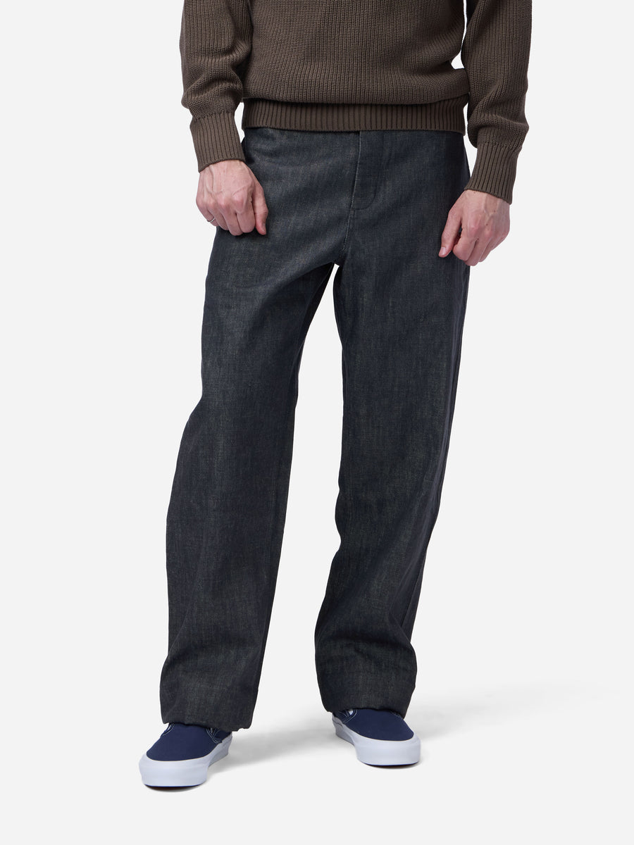 A person wearing dark charcoal gray relaxed-fit denim jeans, a brown ribbed knit sweater, and navy blue sneakers, shown from the chest down against a light gray studio background.