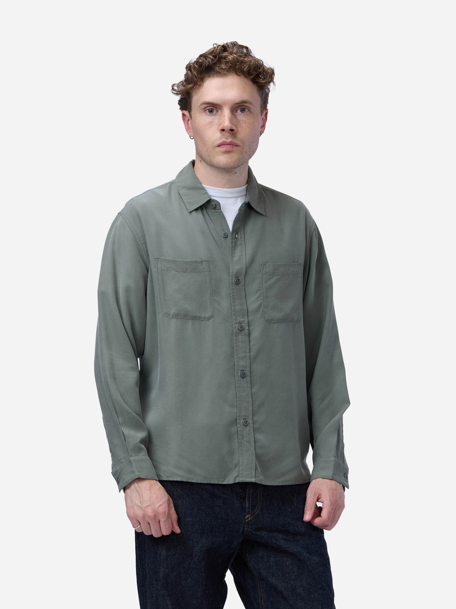 A man wearing a muted sage green long-sleeve button-up shirt over a white undershirt and dark blue jeans, standing against a plain light gray studio background.