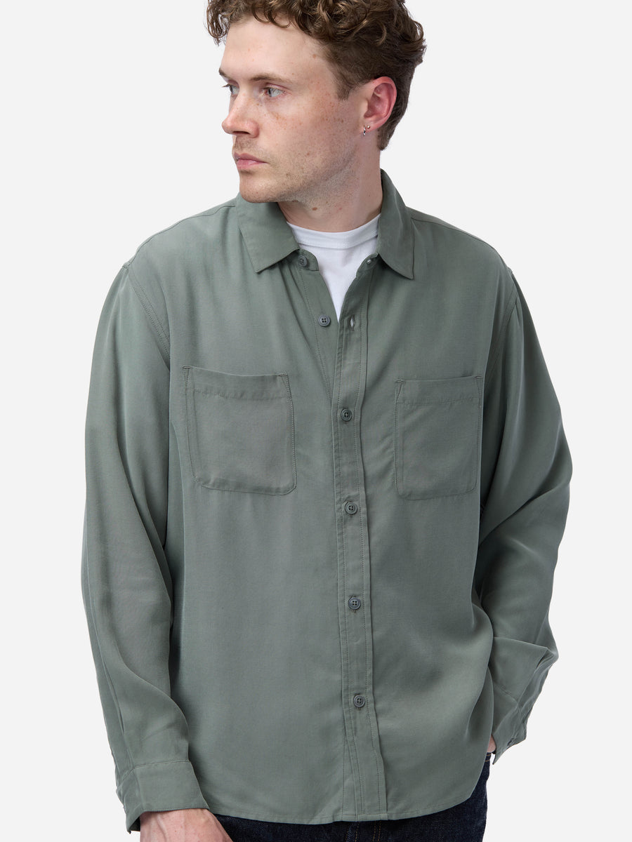 A man wearing a muted sage green long-sleeve button-up shirt over a white t-shirt, photographed against a clean light gray background.