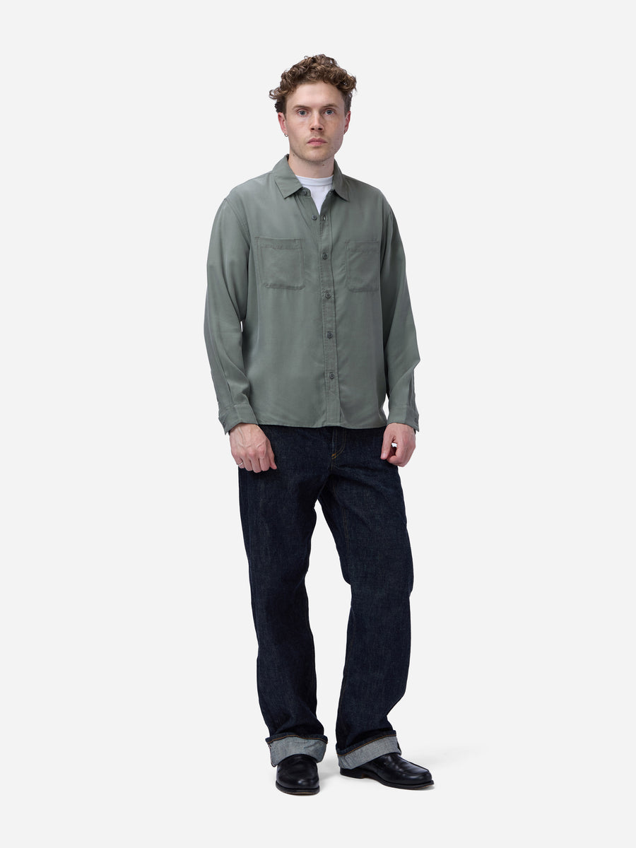 A man wearing a muted sage green long-sleeve button-up shirt over a white undershirt, paired with dark blue jeans and black shoes, standing against a plain light gray background.