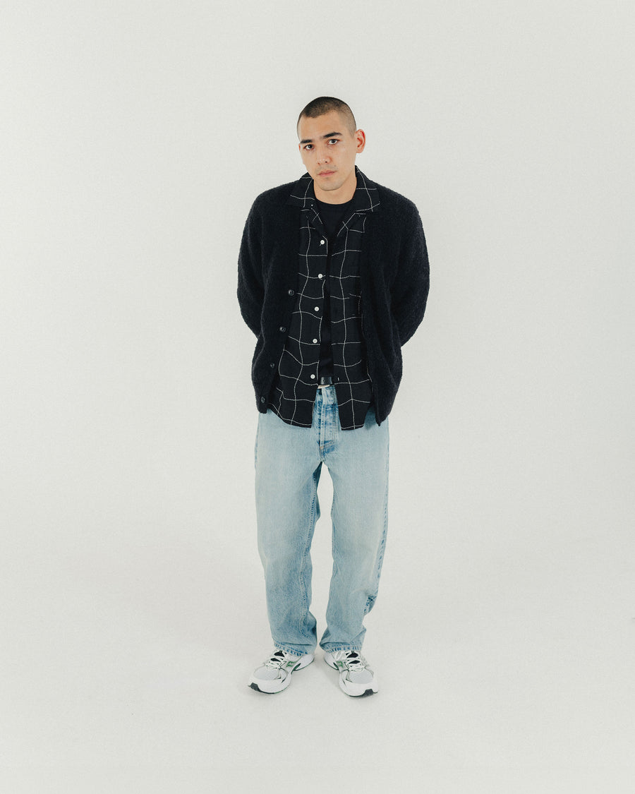 A man in a black button down shirt and light wash blue jeans stands and looks into the camera.