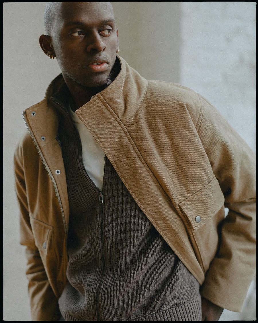 A man in a tan jacket and brown sweater poses.