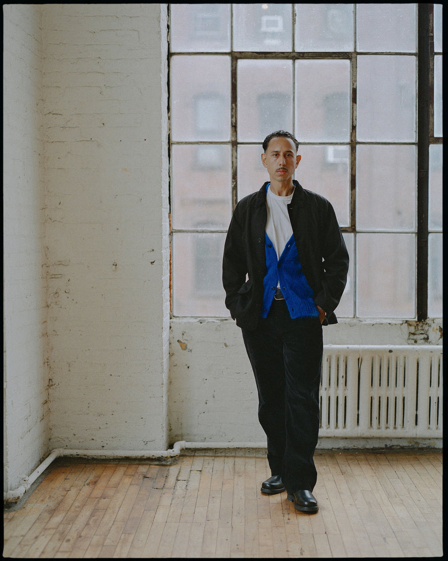 a man in a black jacket and pants over a electric blue cardigan walks into the camera.
