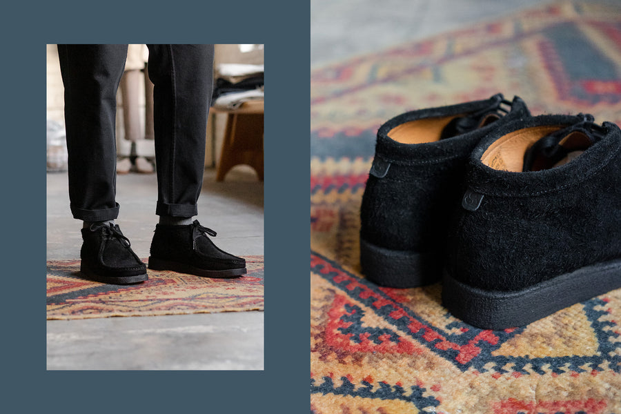 The black suede shoes on feet paired with black tapered pants on a colorful rug.