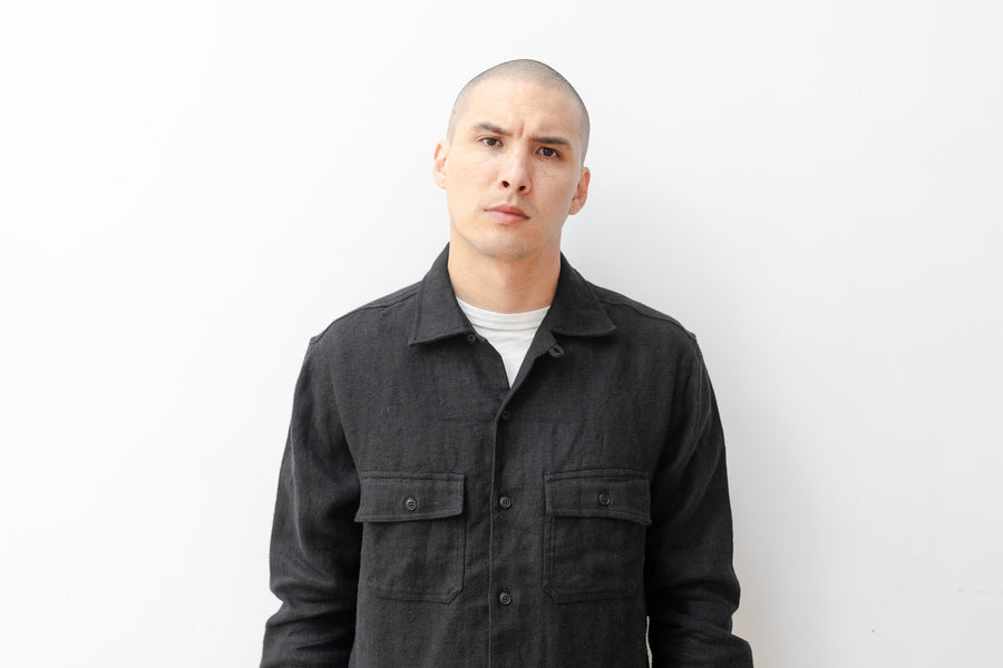 A man in a black button down shirt stands against a white wall.